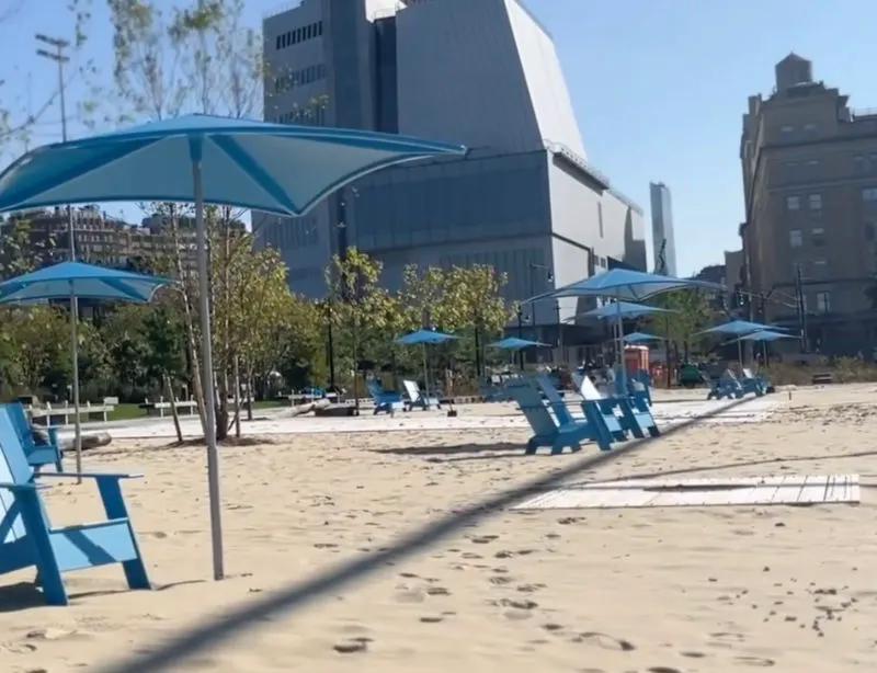 Gansevoort Peninsula, nueva playa urbana de Manhattan en el Meatpacking District. Se ven sillas playeras Adinrondack azules sobre arenas blancas y bajo paraguas de sol azules y de fondo se distingue el edificio del Museo Whitney de Arte Americano. Captura de pantalla del video de presentación de la atracción disponible en la página del Hudson River Park https://hudsonriverpark.org/locations/gansevoort-peninsula/