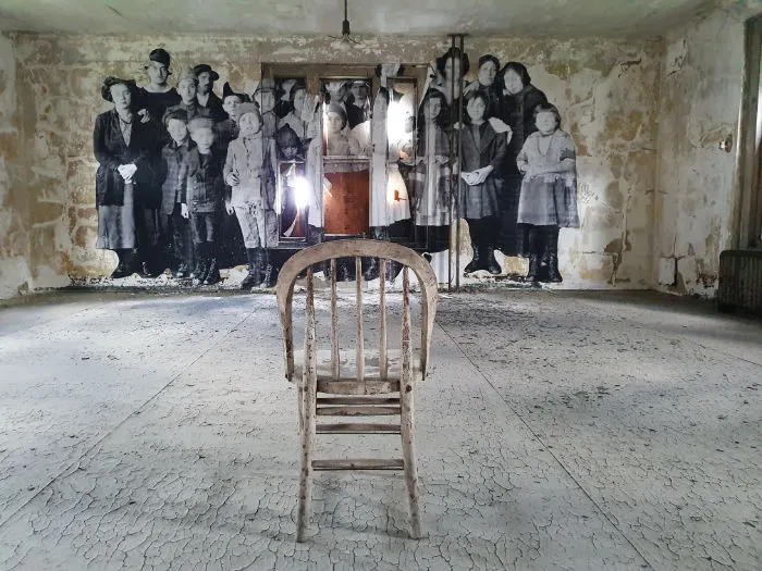 Interior de una de las salas del Hospital de Inmigrantes de Ellis Island. Foto de Andrea Hoare Madrid durante el recorrido del Hard Hat Tour. Se ve una antigua silla vacía en una sala abandonada y sin restaurar, en el fondo una gigantografía de una foto de un grupo de inmigrantes que pasaron por el Hospital.