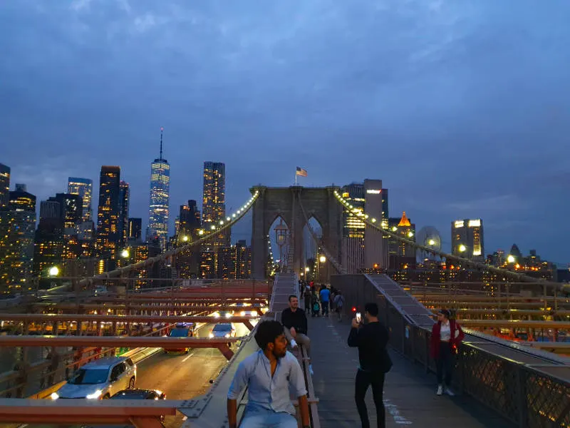 Personas sacando fotos en el Brooklyn Bridge de noche. Al fondo se ven los rascacielos de Lower Manhattan con sus luces encendidas. Foto de Andrea Hoare Madrid