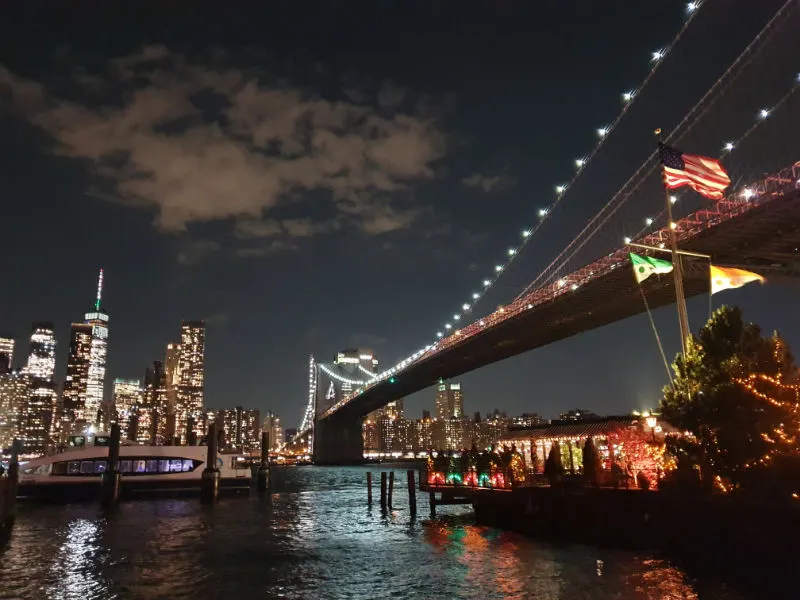 Photograph by Andrea Hoare Madrid of the Brooklyn Bridge lit up on the right and the skyscrapers of Lower Manhattan on the left. Image taken from Brooklyn Bridge Park in DUMBO.