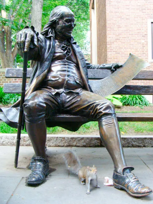 Estatua de de bronce de Benjamin Franklin sentada en una banca de la Universidad de Pensilvania en Filadelfia, a los pies de la estatua se ve una ardilla. Foto de Andrea Hoare Madrid